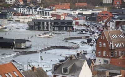   Fotoet er et luftfoto, der viser en stormflod der holdes ude af stormflodsvægge ved havnen i Lemvig. Vandet er nået til toppen af stormflodsvæggene og er på vej til at oversvømme den vej, der grænser op til havneområdet. 