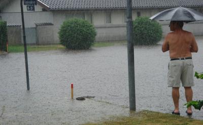   Fotoet viser en villavej, der er helt oversvømmet af vand. Det regner fortsat kraftigt. I højre hjørne ses en mand med en paraply, han står og kigger ud over vandmasserne.