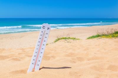 Fotoet viser en strand med sand, blåt vand og blå himmel. I sandet står et termometer som viser 35 ºC