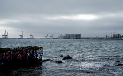   Fotoet viser et havneområde med lastekraner og høje beboelseshuse i baggrunden. I forgrunden ud til havnen ses en kajkant og store sten i vandet. Det er overskyet og gråt.
