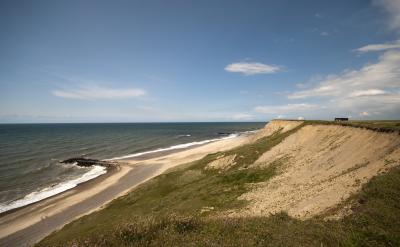   Fotoet viser et kystområde med en strand, hvor havet skyller roligt ind. Fra stranden er der en høj skrænt op mod det bagvedliggende naturområde. Himlen er blå med lette hvide skyer