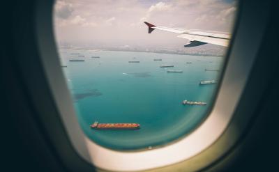   Fotoet viser udsigten ud af et flyvindue. Gennem vinduet ses havet med en masse containerskibe. Længere ude ses kysten med en storby