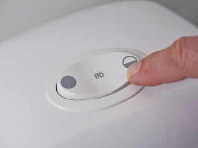 Fotoet viser toppen af et to-skyls toilet og en persons finger, der trykker på det lille skyl med 3 liter.